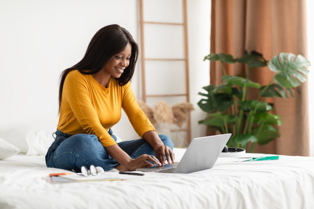 Black Young Woman Using Laptop Typing Sitting In Bedroom Indoors