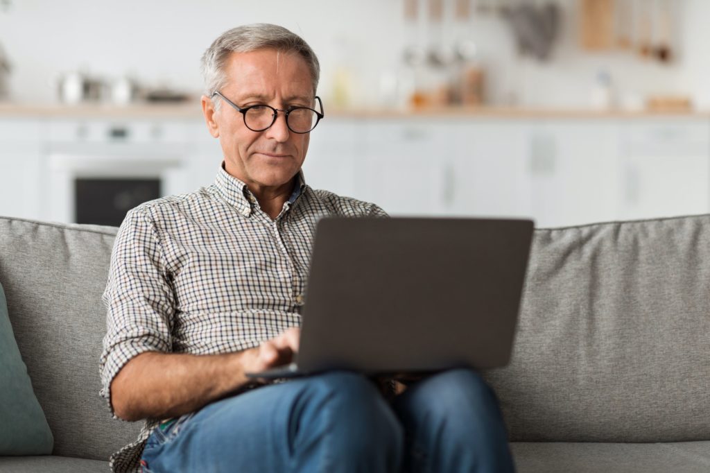 Senior Male Using Laptop Sitting On Couch At Home